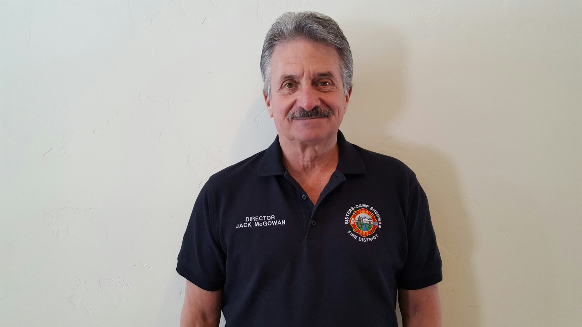 A man in a navy polo shirt with a fire department logo and text "Director Jack McGowan" stands against a plain wall.