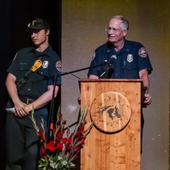 Two uniformed men, one at a podium with a horse emblem and flowers, the other standing beside, possibly at a ceremony or event.