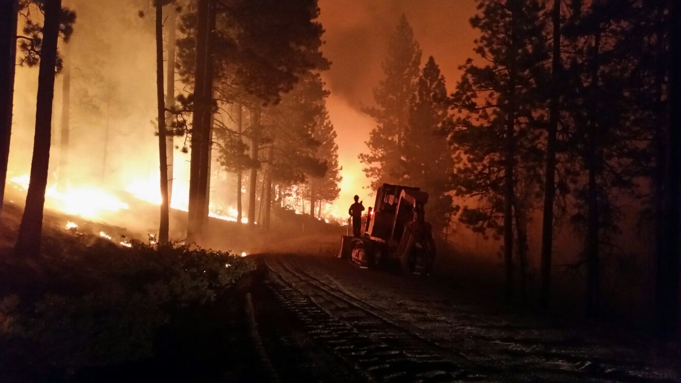 A forest scene with intense fire, smoke, and silhouetted trees. A person stands beside heavy machinery.