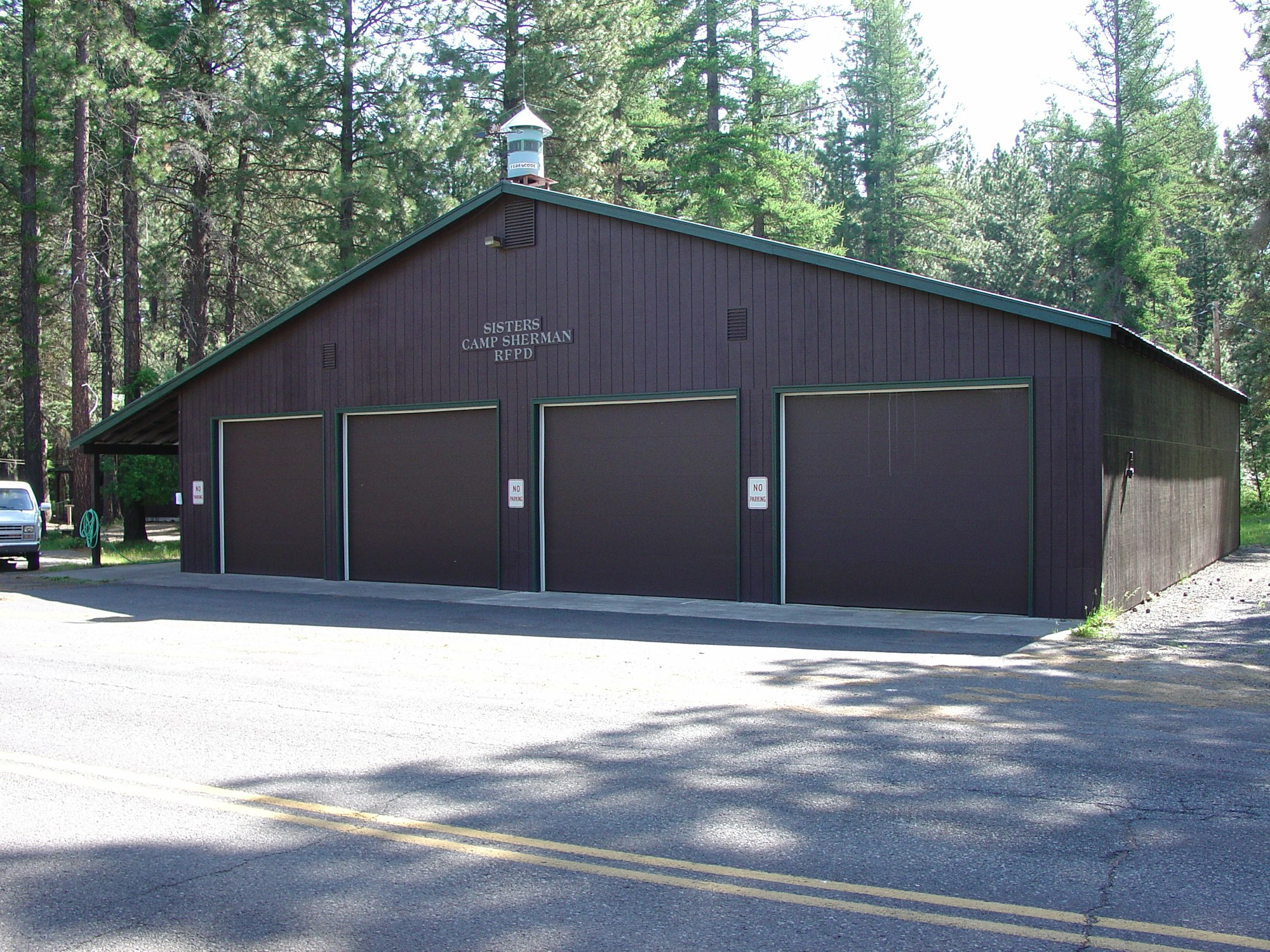 A brown building with three garage doors, labeled "Sisters Camp Sherman RFPD," surrounded by trees.