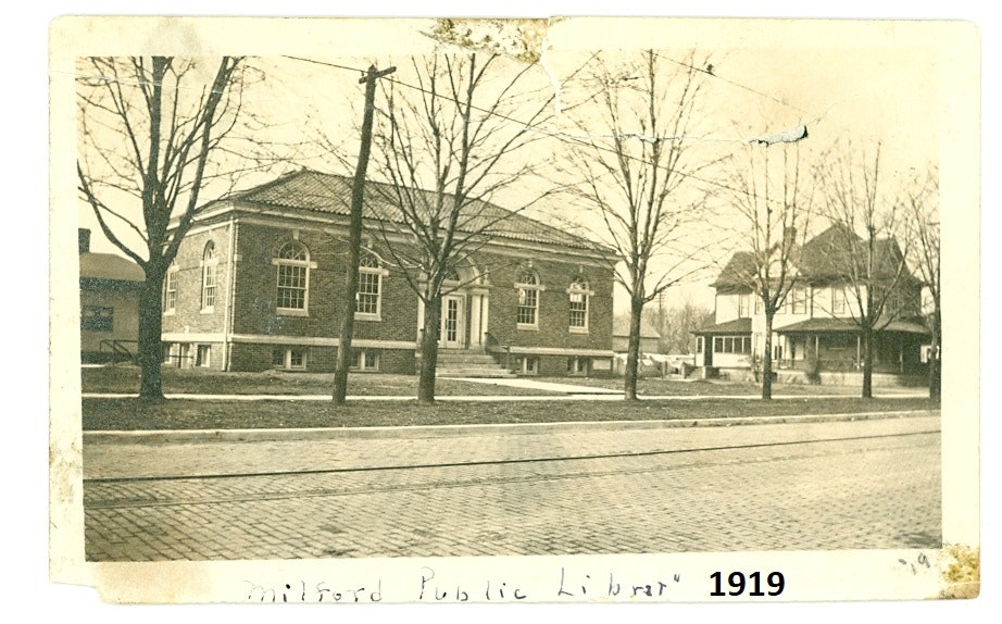 Historical photo of Milford Public Library from 1919, featuring a brick building and surrounding trees.