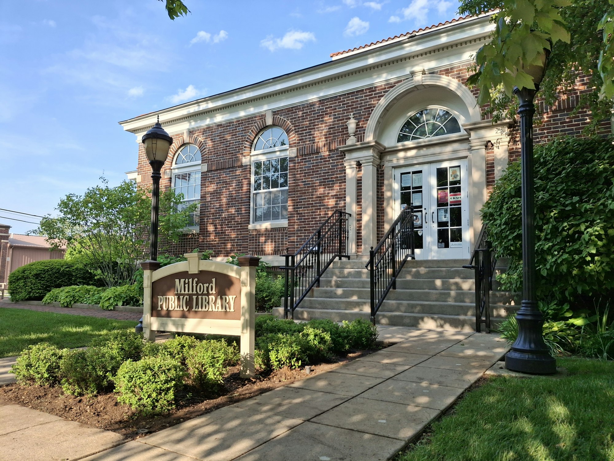 A view of the Milford Public Library, featuring its brick facade, steps, greenery, and a welcoming sign.