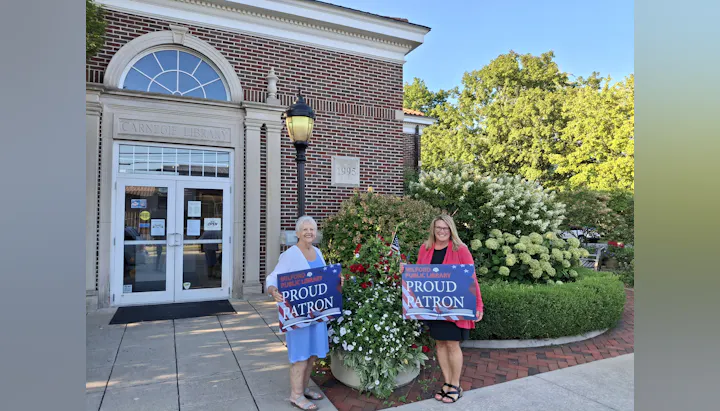 Two women stand outside the Carnegie Library holding signs that say "Proud Patron" of the Milford Public Library.