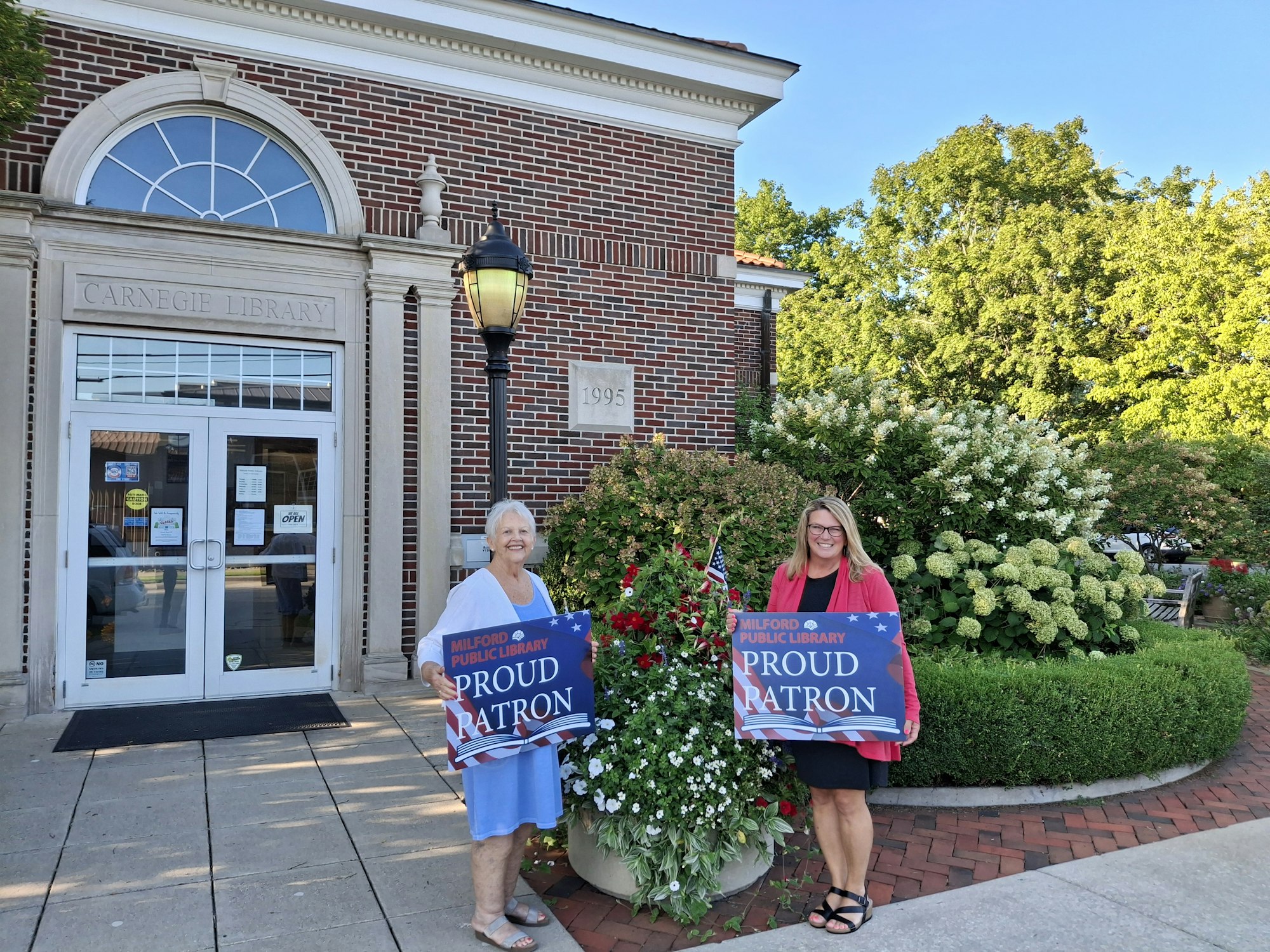 Two women stand outside the Carnegie Library holding signs that say "Proud Patron" of the Milford Public Library.