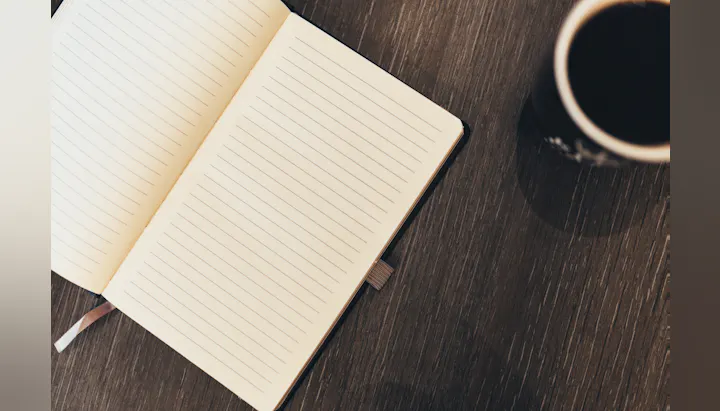 An open lined notebook on a wooden surface next to a cup of coffee. Ideal for writing thoughts or notes.