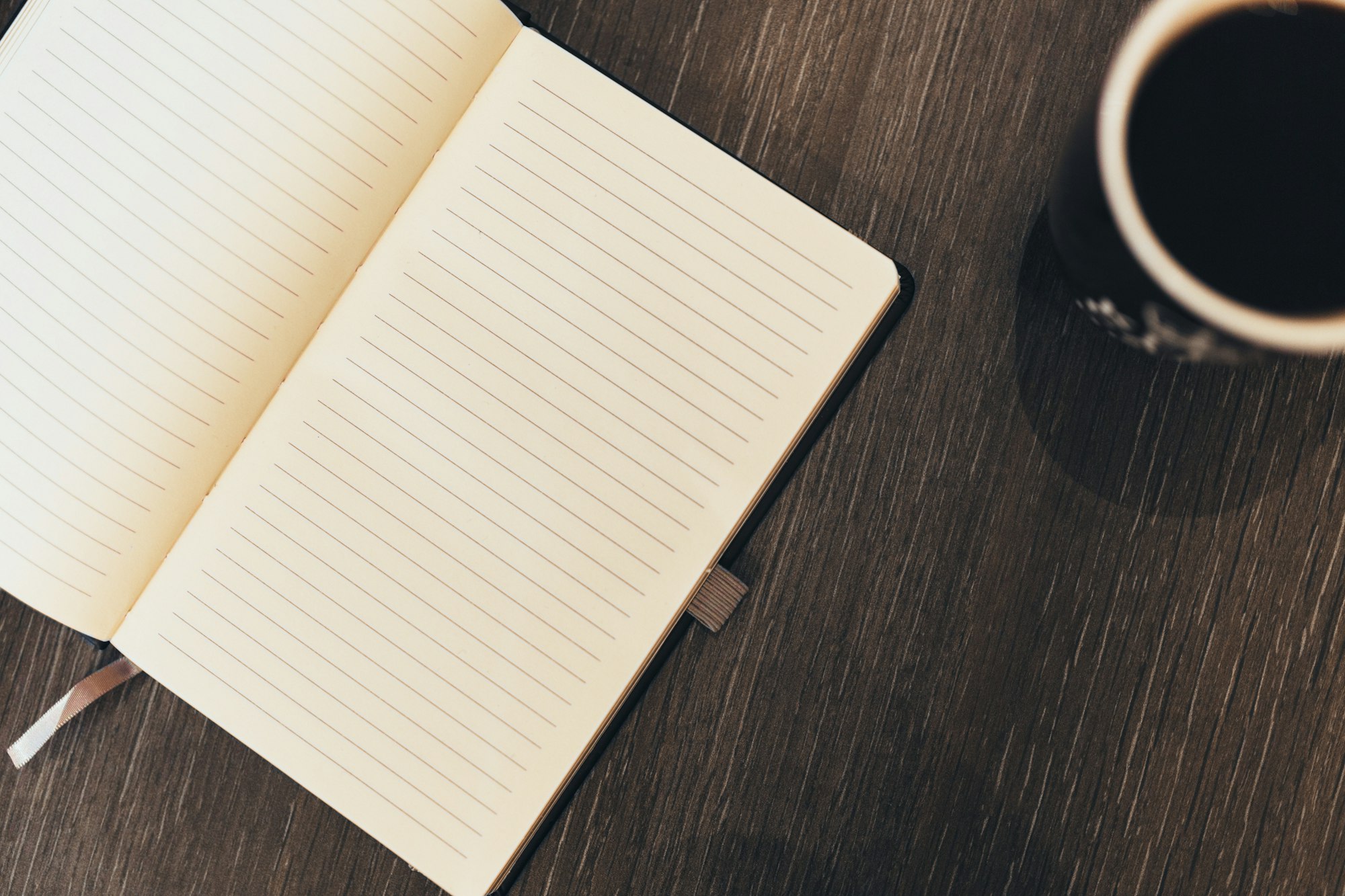 An open lined notebook on a wooden surface next to a cup of coffee. Ideal for writing thoughts or notes.