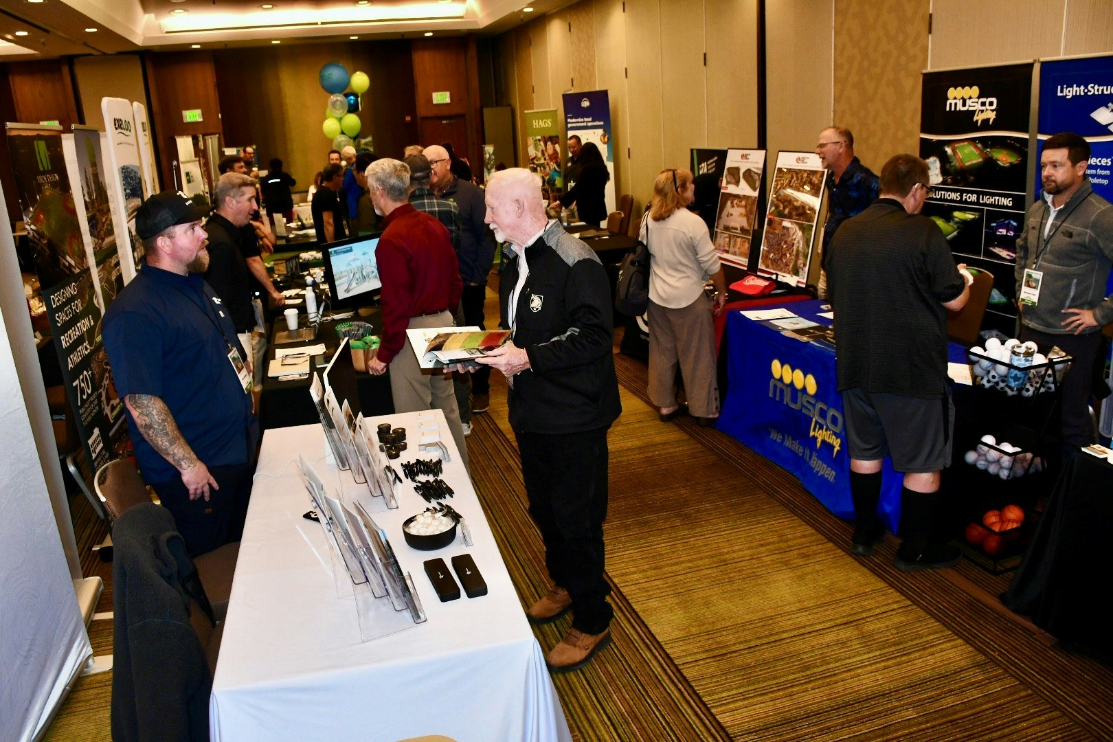 A busy trade show with various booths displaying products and services related to recreational facilities, lighting, and design.