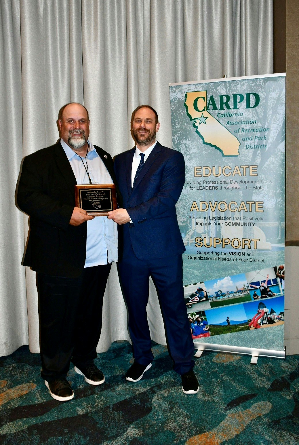 Two men are posing with an award in front of a banner for the California Association of Recreation and Park Districts (CARPD).