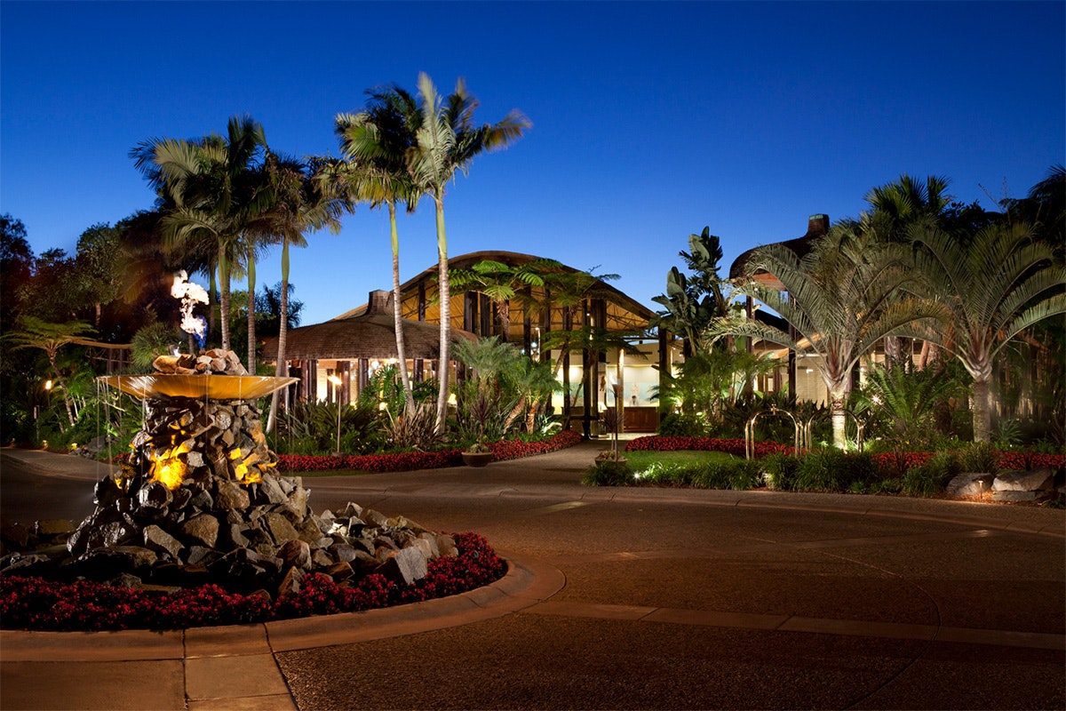 A tropical resort entrance at dusk with lit torches, palm trees, and a welcoming pathway.