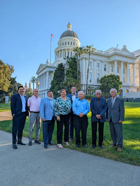 A group of eight individuals poses in front of a large white building with a dome, likely a government structure.