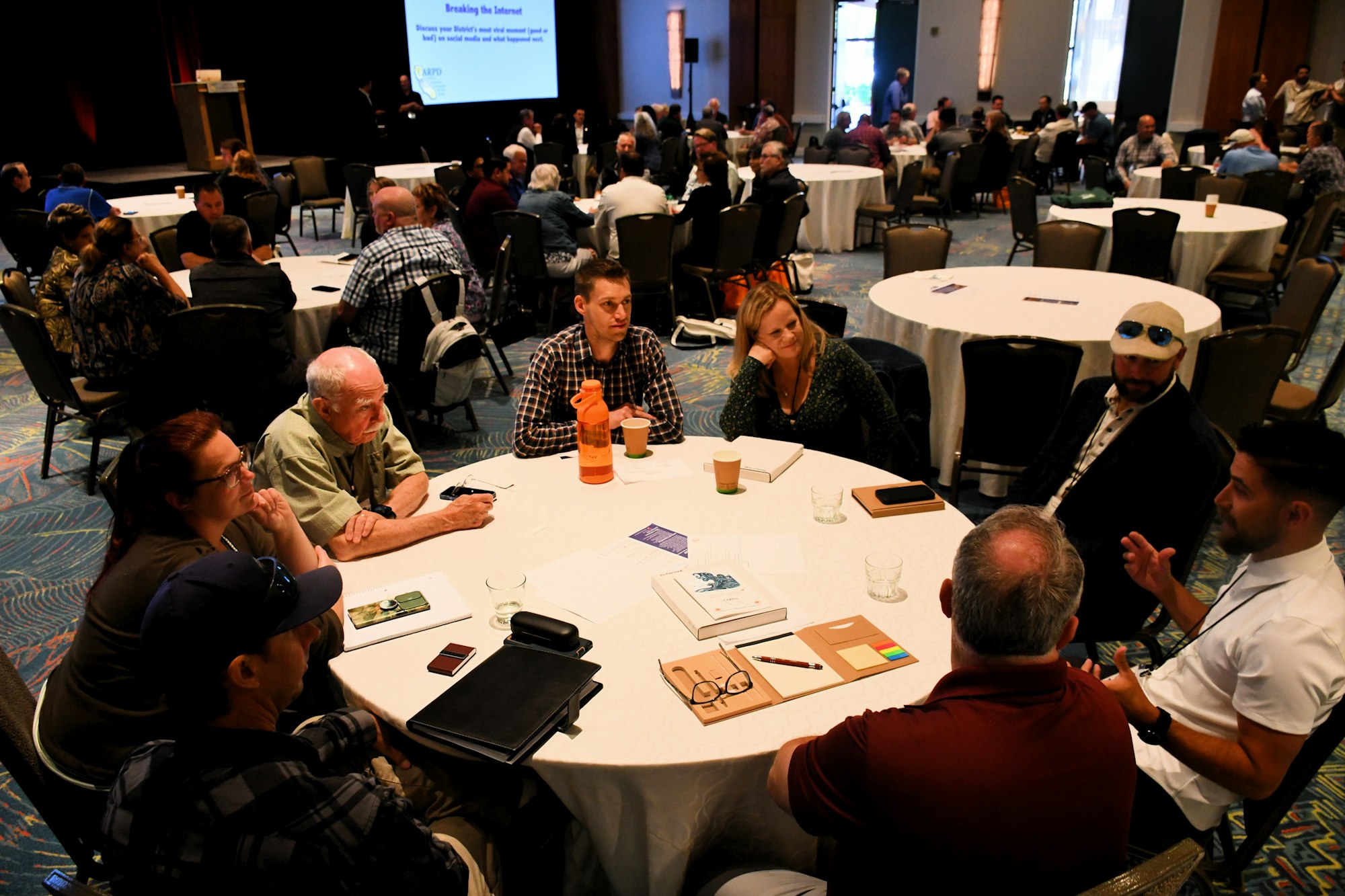 A group of people sit at round tables during a conference, engaged in discussion with notebooks and drinks on the tables.