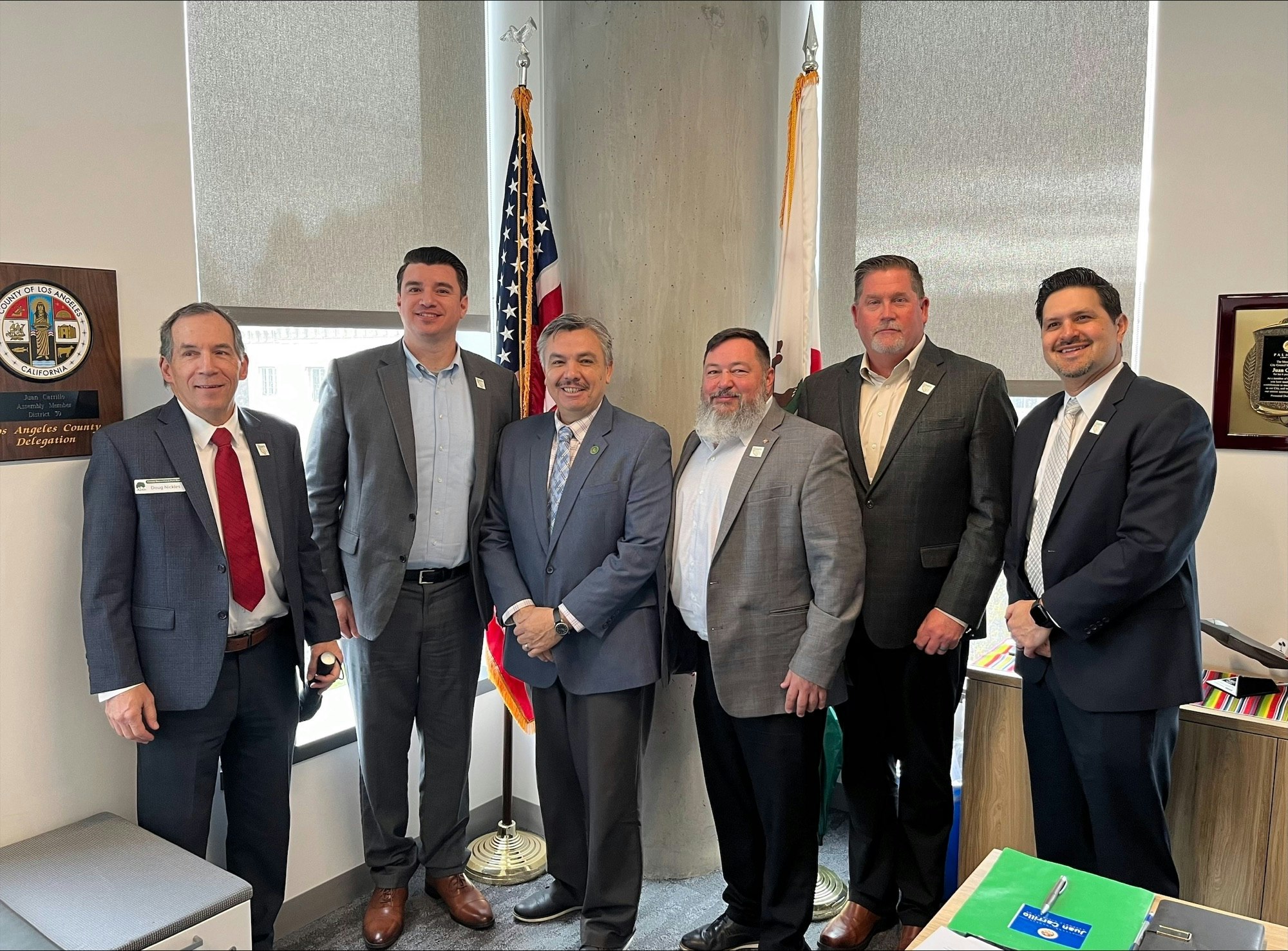 Six men in suits stand together in an office with flags and plaques in the background.