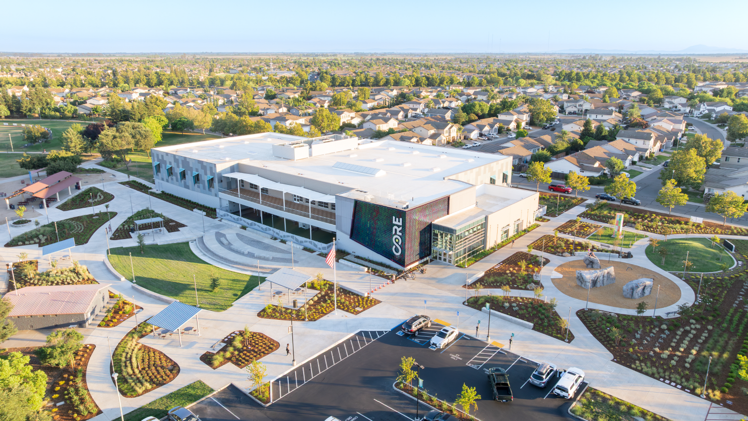 A modern building surrounded by landscaped areas, parking, and residential neighborhoods; features paths and playgrounds.