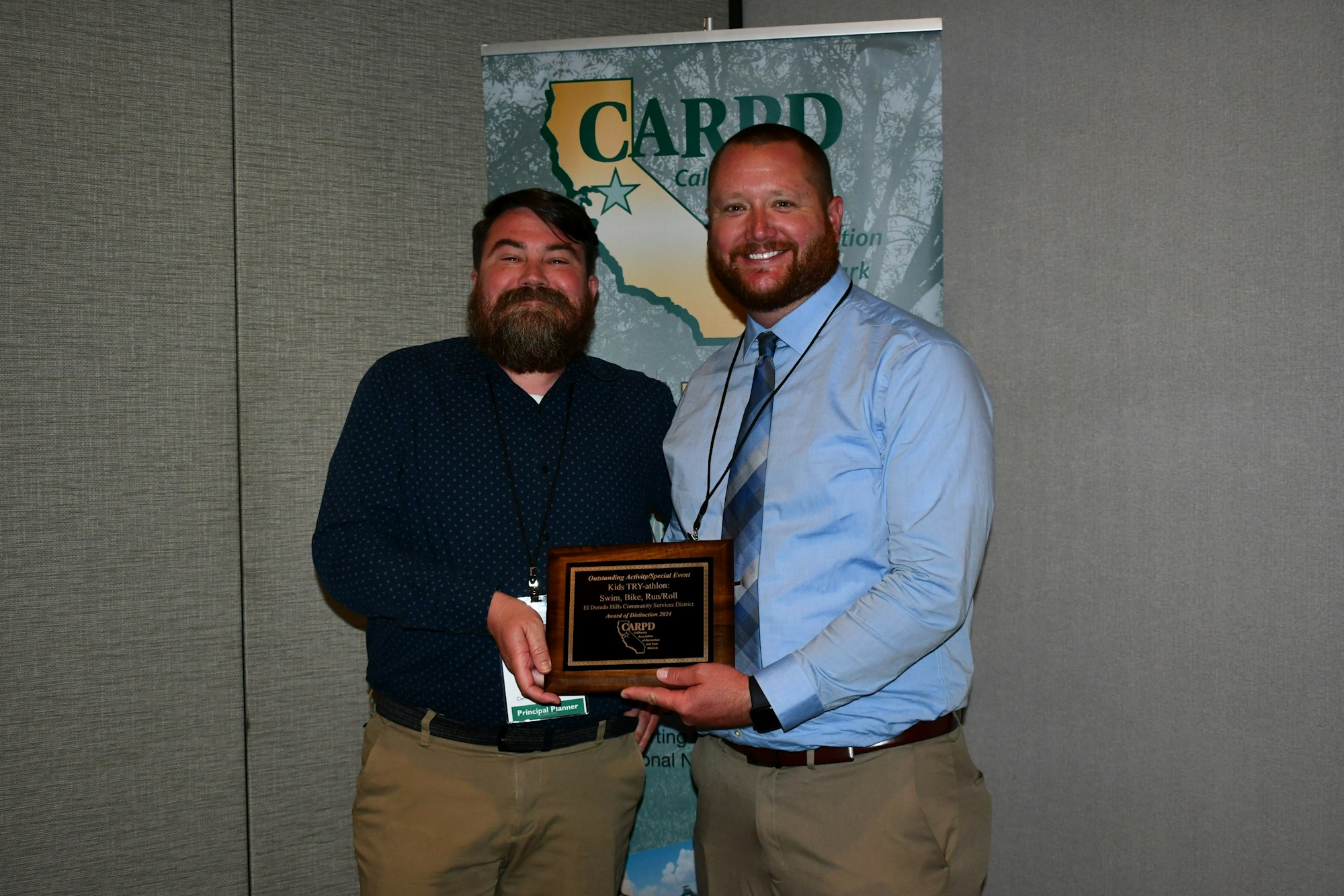Two men smiling, one holding an award, in front of a banner.