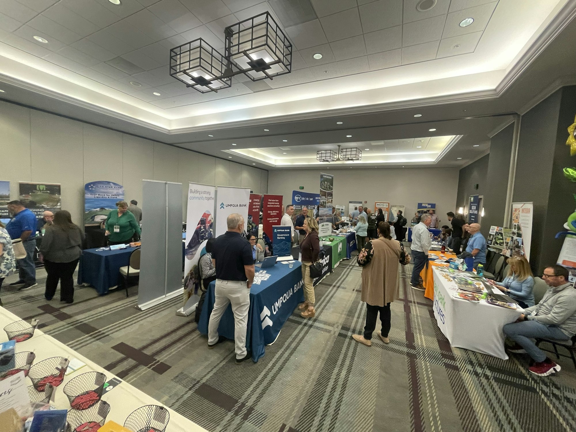 An indoor trade show or expo with booths, people networking, and various company banners.