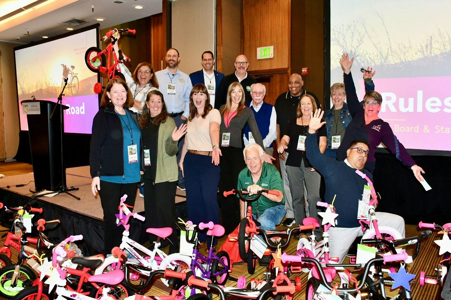 A group of people poses enthusiastically in front of a stage, surrounded by colorful children's bicycles.
