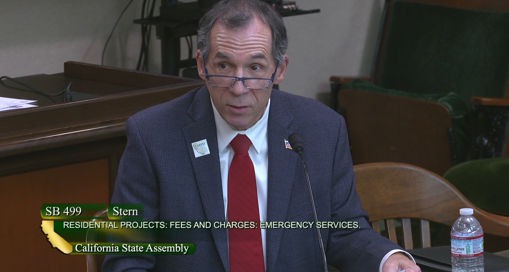A man wearing glasses and a red tie speaks into a microphone at the California State Assembly about SB 499.