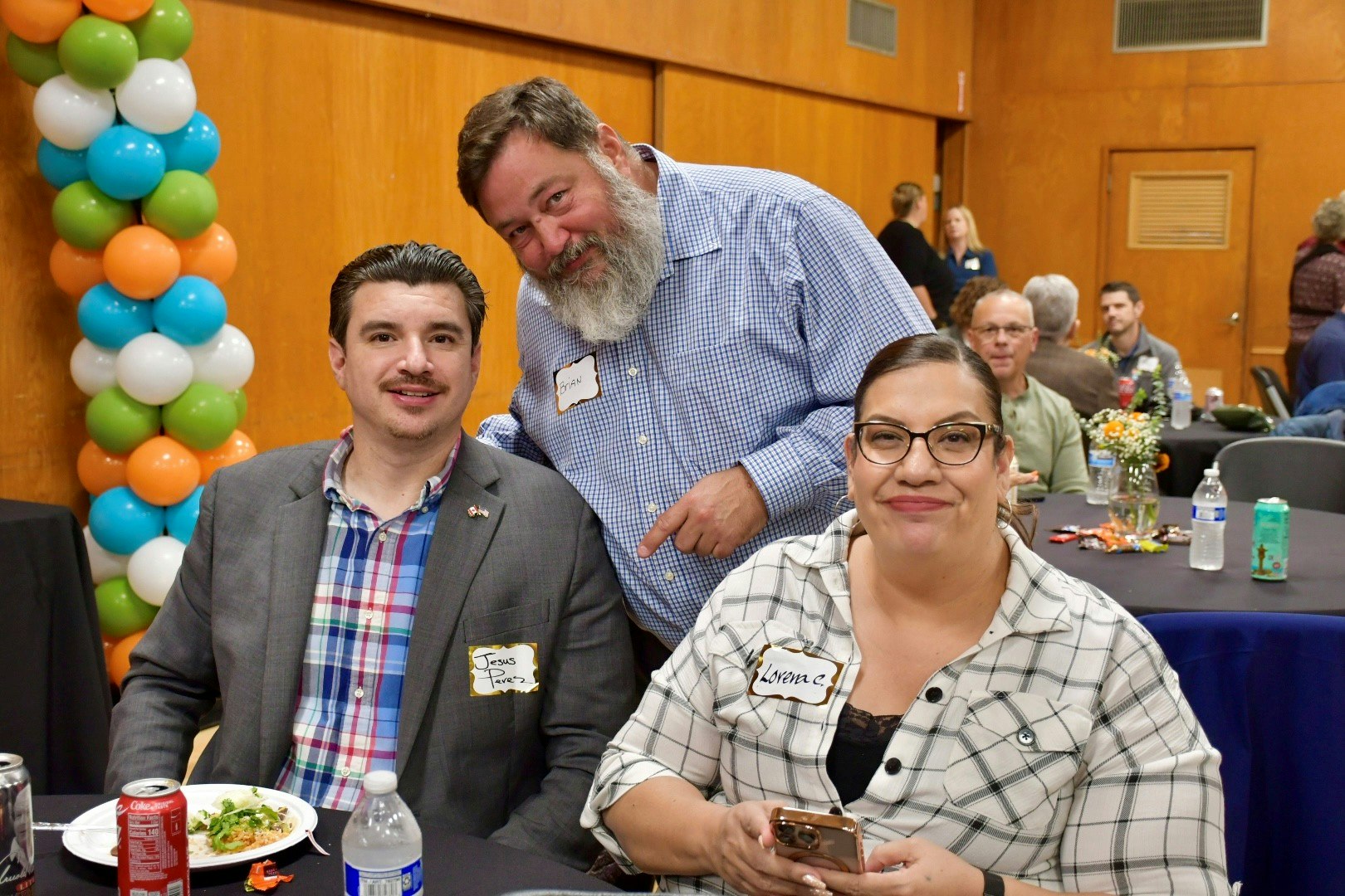 A group of three people smiles for a photo at an event decorated with colorful balloons, with food and drinks on their table.