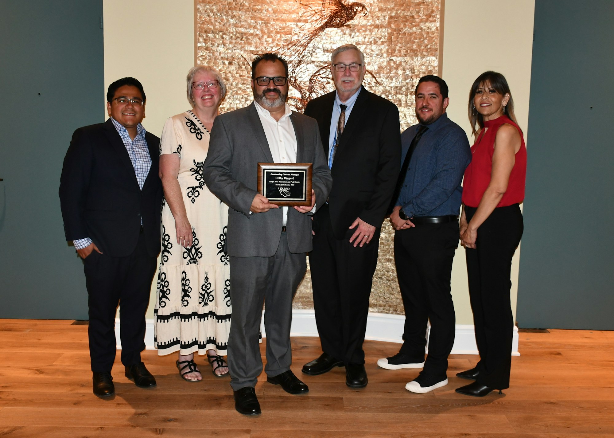 Seven people posing, one holding an award plaque, standing in front of a textured wall.