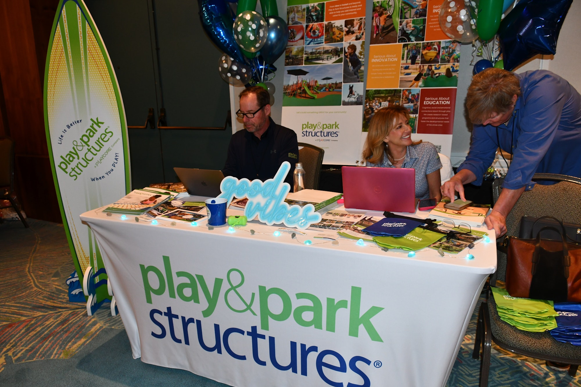People at a "play & park structures" booth with laptops, brochures, and a "good vibes" sign.