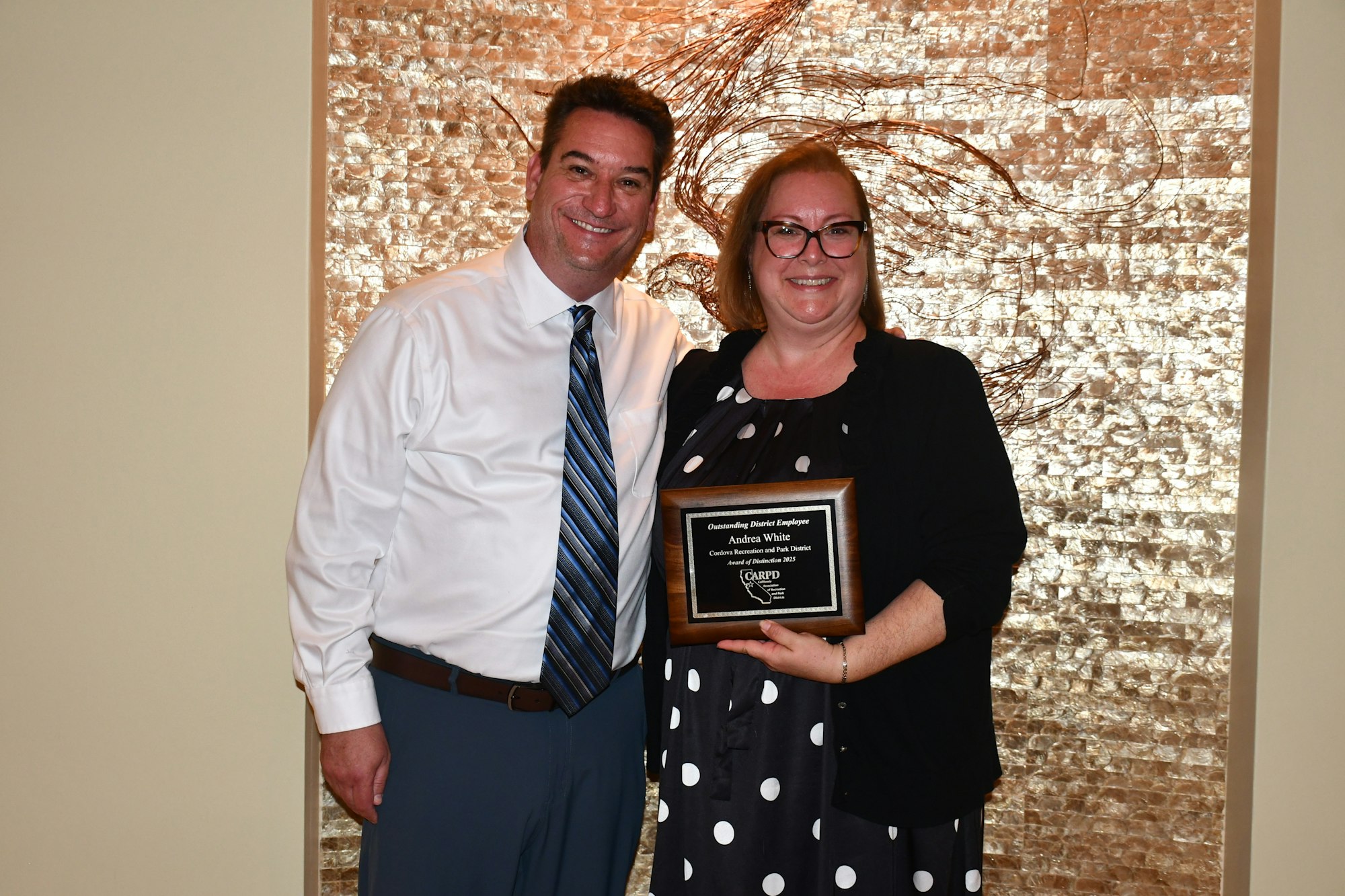 Two people smiling, one holding a plaque for "Outstanding District Employee" award, standing against a textured wall backdrop.