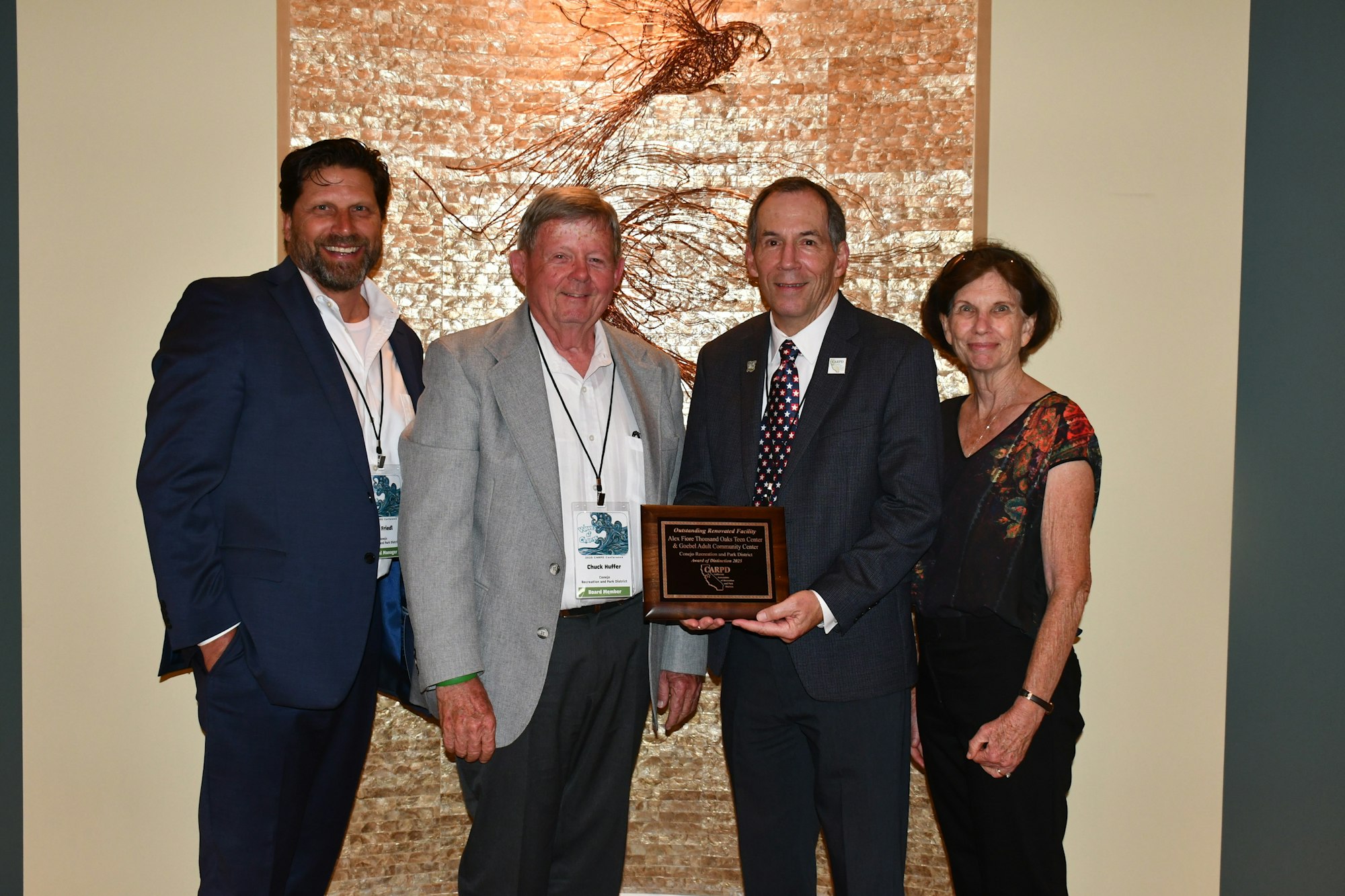 Four people posing with a plaque, standing in front of a textured wall art piece.