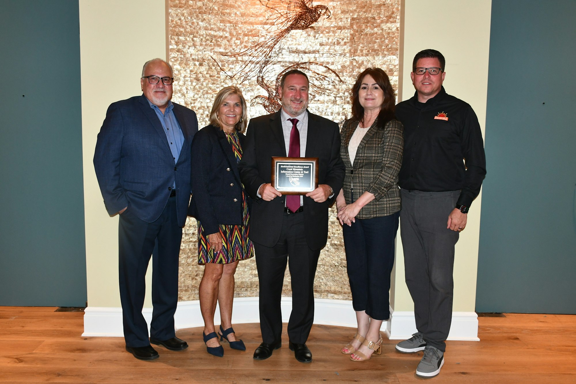 Five people pose in front of artwork. The person in the middle holds a plaque.
