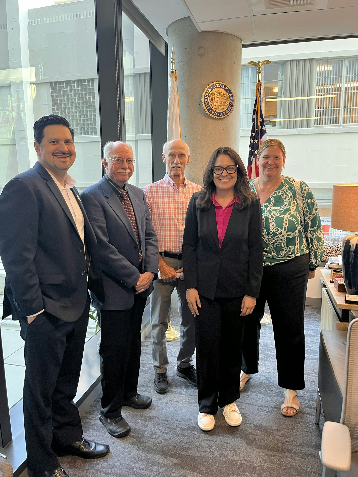 A group of five adults, dressed in business attire, pose for a photo in an office setting with flags and a state seal in view.
