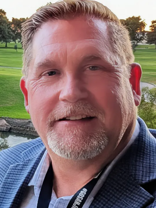 A close-up portrait of a man smiling, dressed in a blazer, with a scenic green background of trees and a golf course.