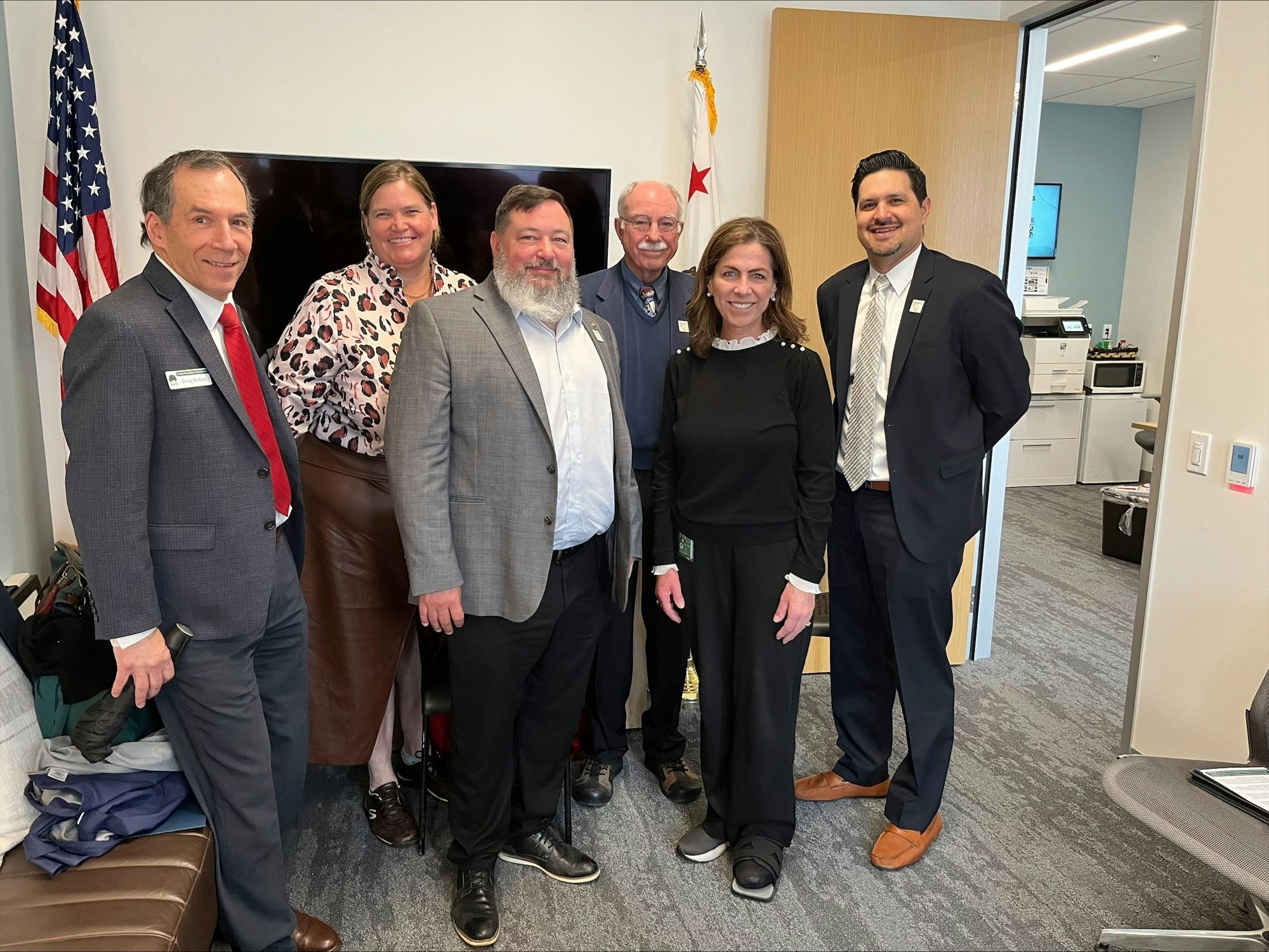 A group of six people in business attire posing for a photo in an office setting with US and state flags.