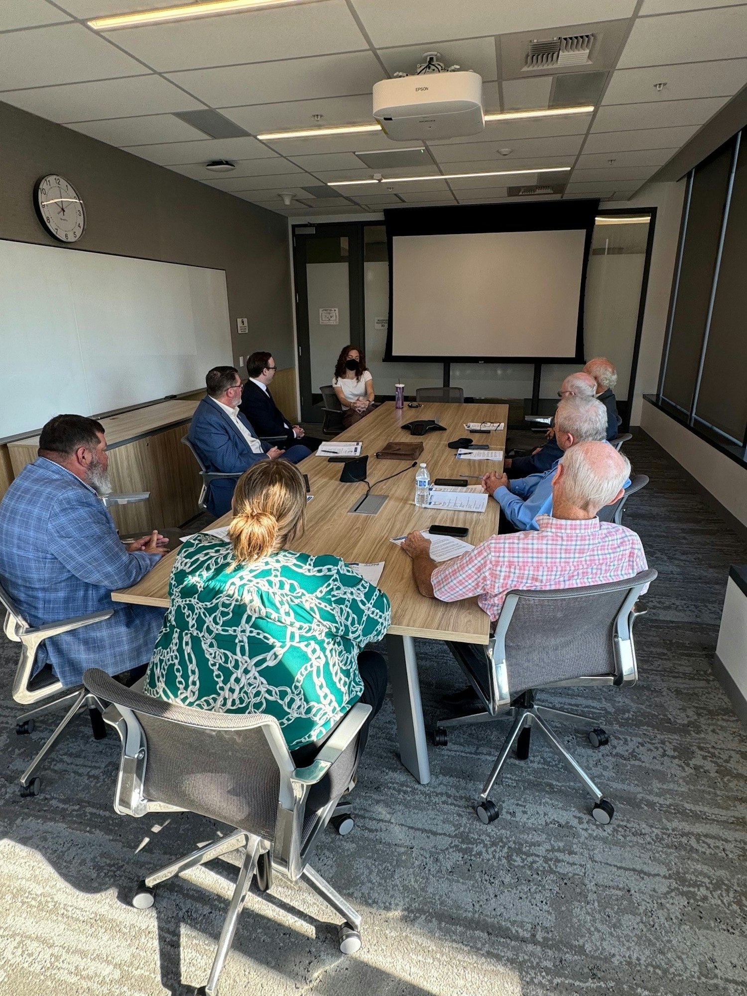 A group of people in business attire seated around a conference table, engaged in a meeting, with a projector screen in the background.