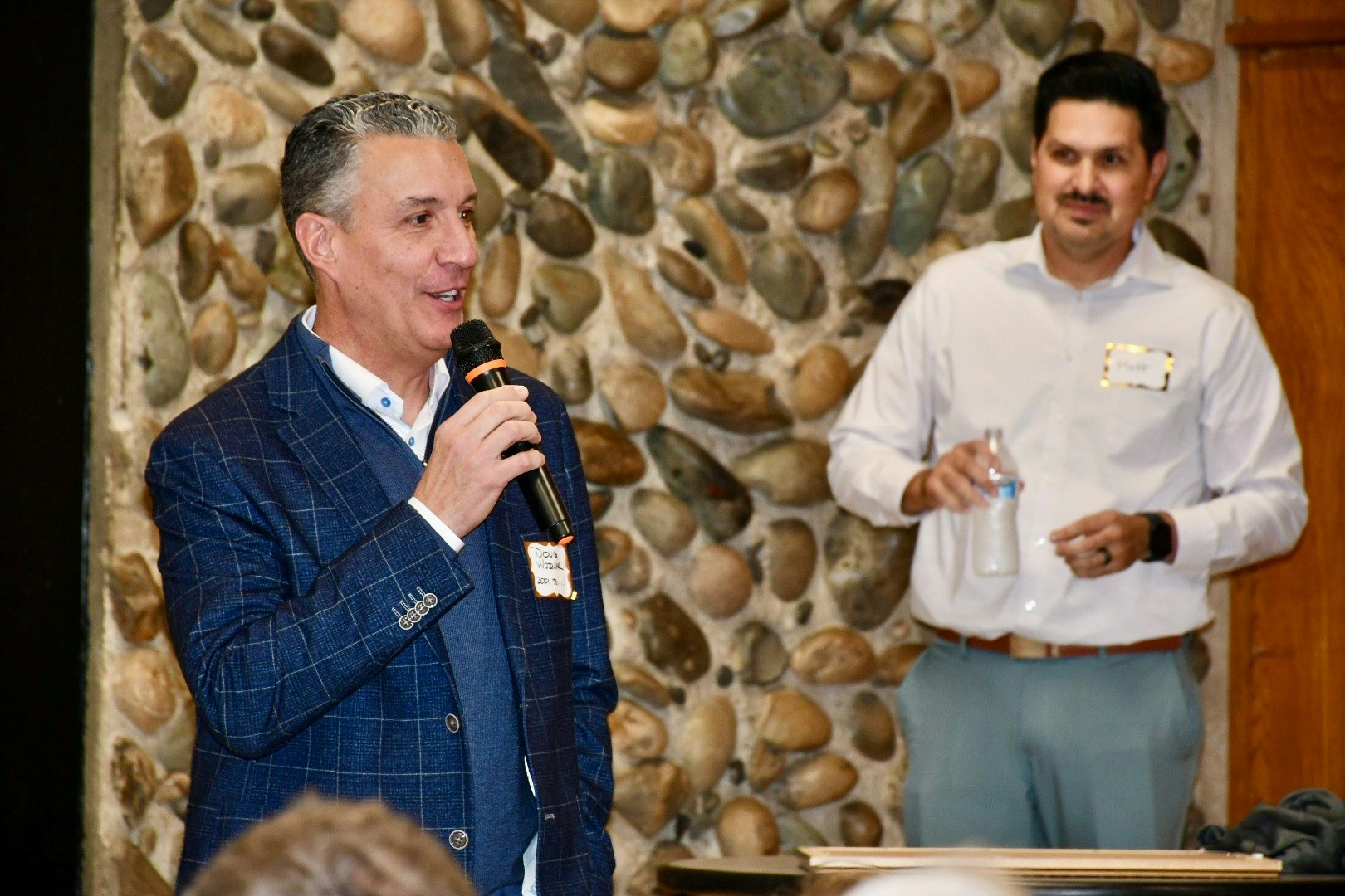 Two men are speaking at an event; one is holding a microphone, while the other holds a water bottle. A stone wall is in the background.