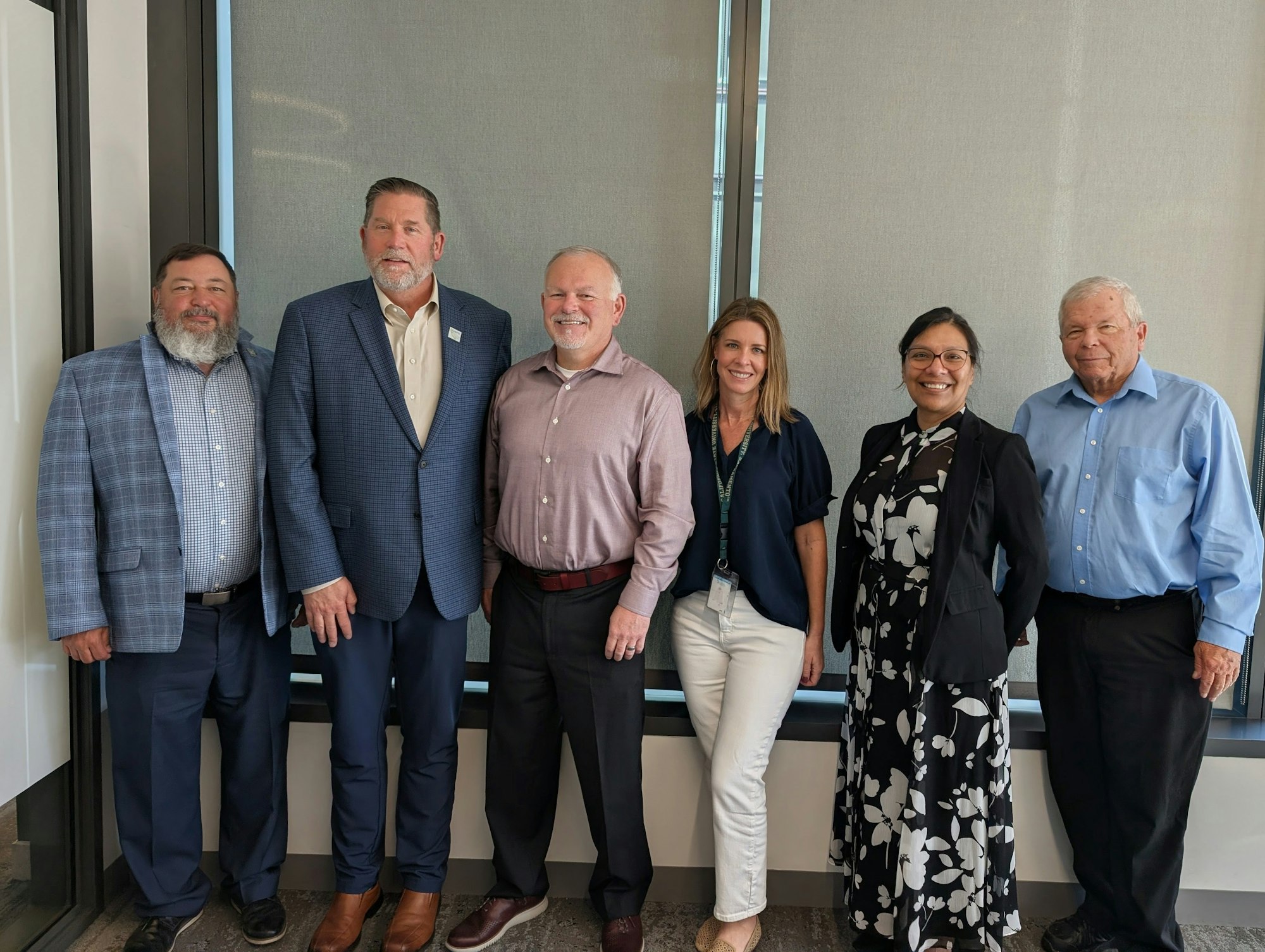 A group of six people stands in front of a window, all dressed in professional attire, smiling for a photo.