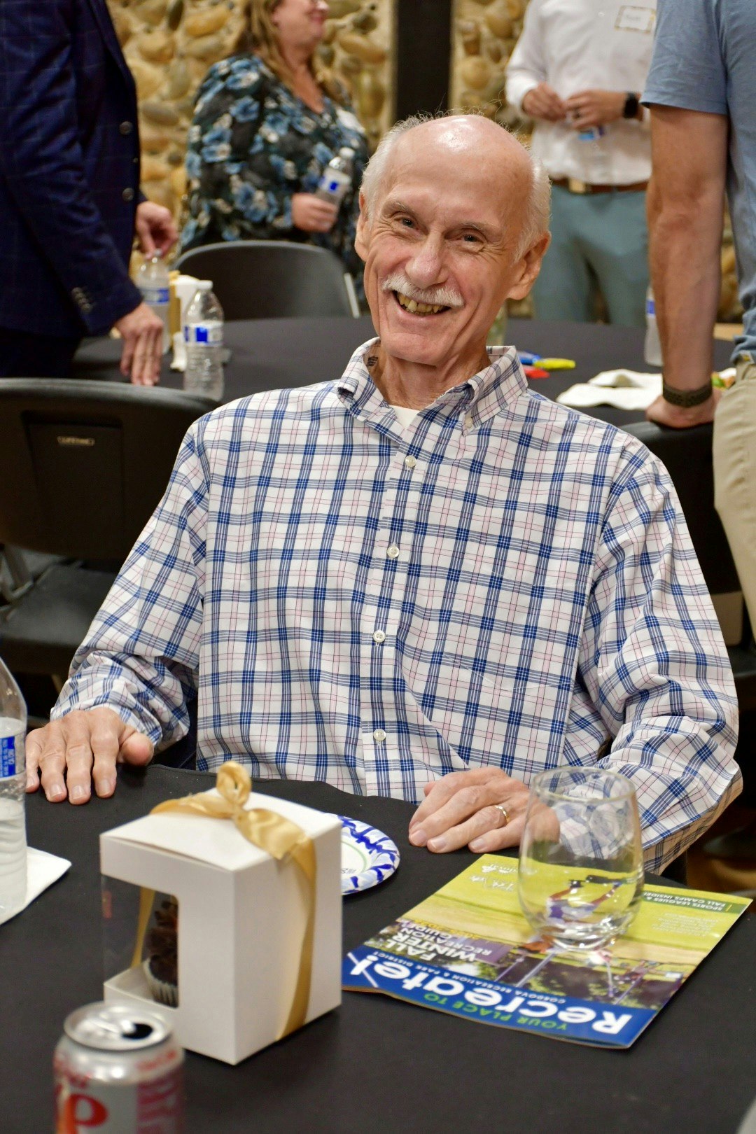 A smiling older man in a plaid shirt sits at a table with drinks and a gift box, enjoying a social event.
