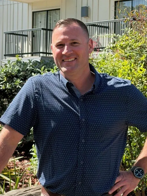 A smiling man in a blue shirt and khakis poses by a wooden railing with greenery and a building in the background.