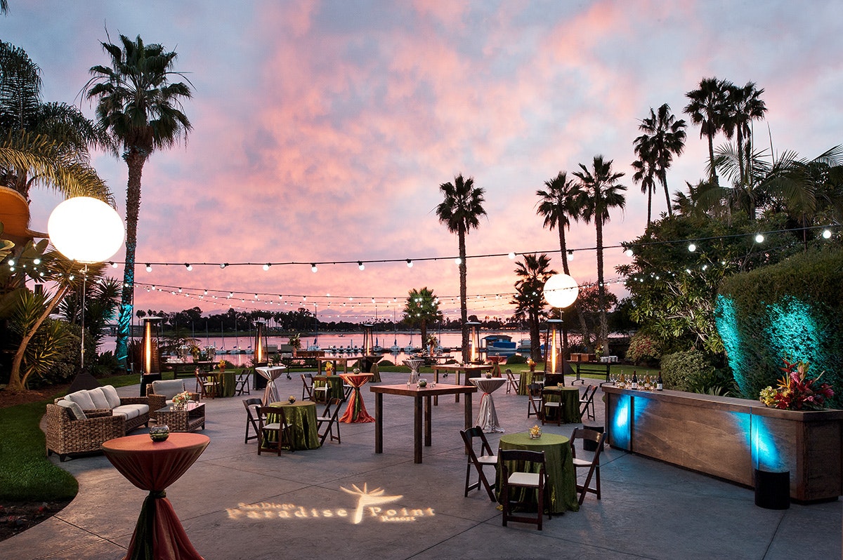 Outdoor seating area at dusk with string lights, palm trees, a bar, and a colorful sky.