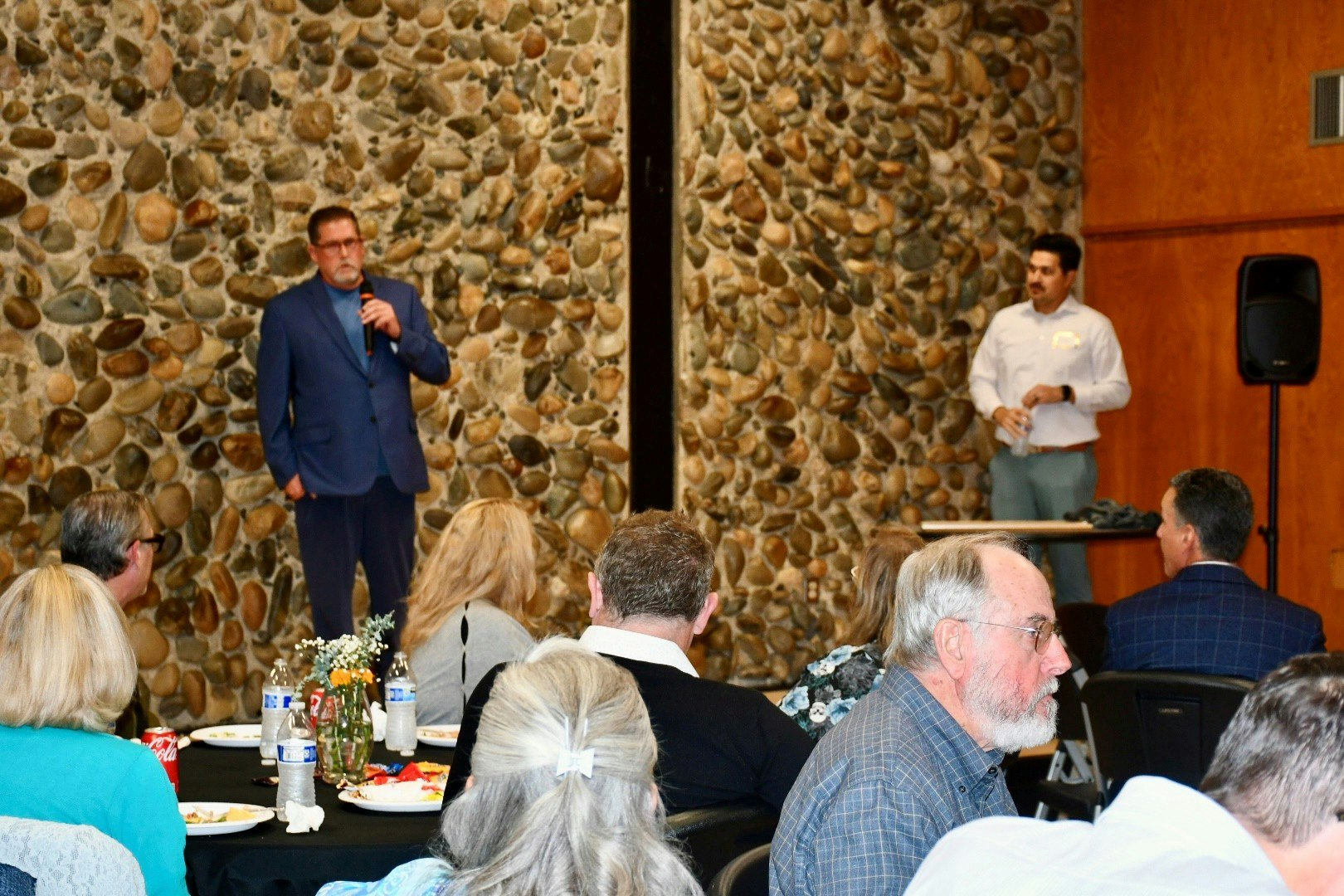 An event with attendees sitting at tables, while two speakers engage with the audience against a stone wall backdrop.