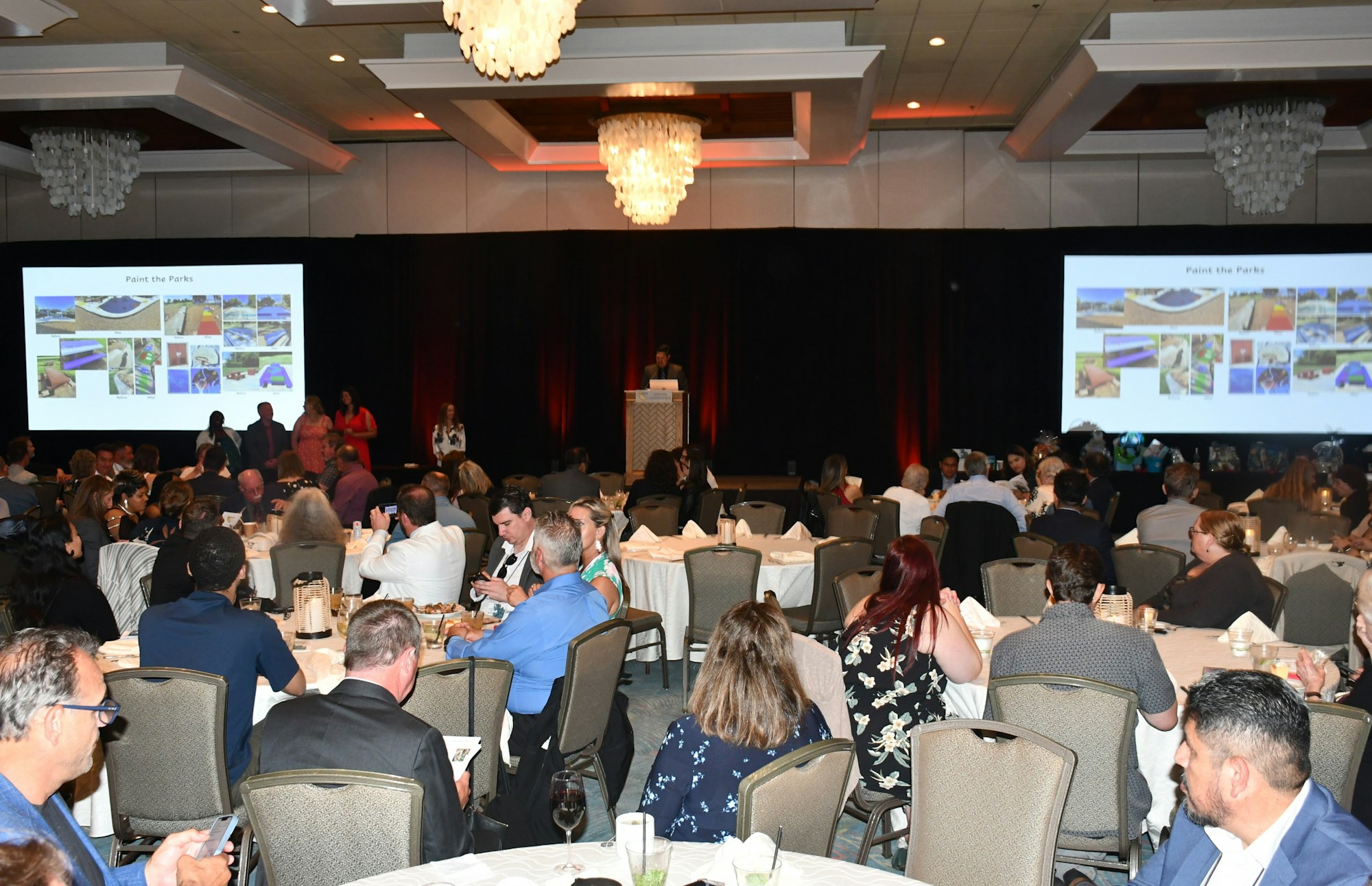 A conference room with people seated at tables, watching a presentation titled "Paint the Parks" on two screens.