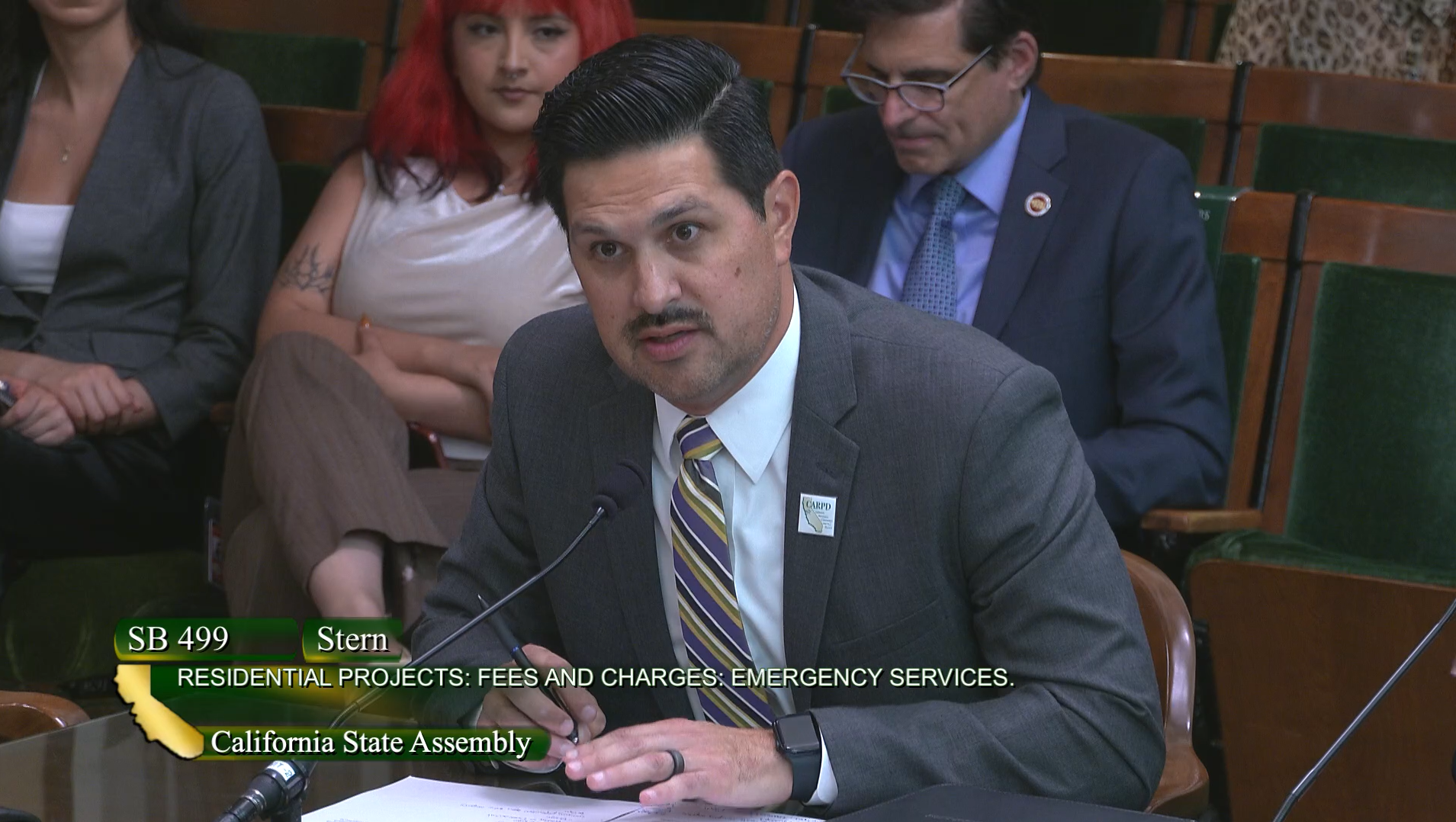 A man in a suit speaking at a California State Assembly meeting about SB 499, with people seated behind him.