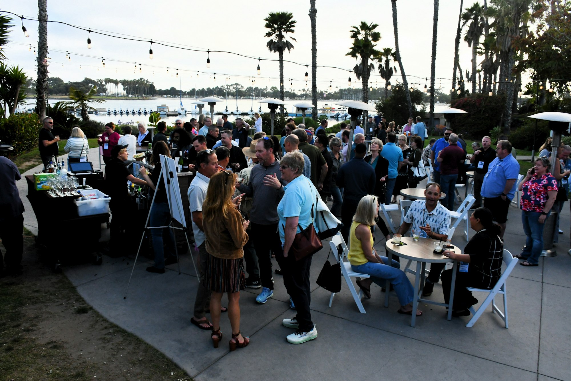 A group of people socializing outdoors near a waterfront, with tables, chairs, and string lights overhead.
