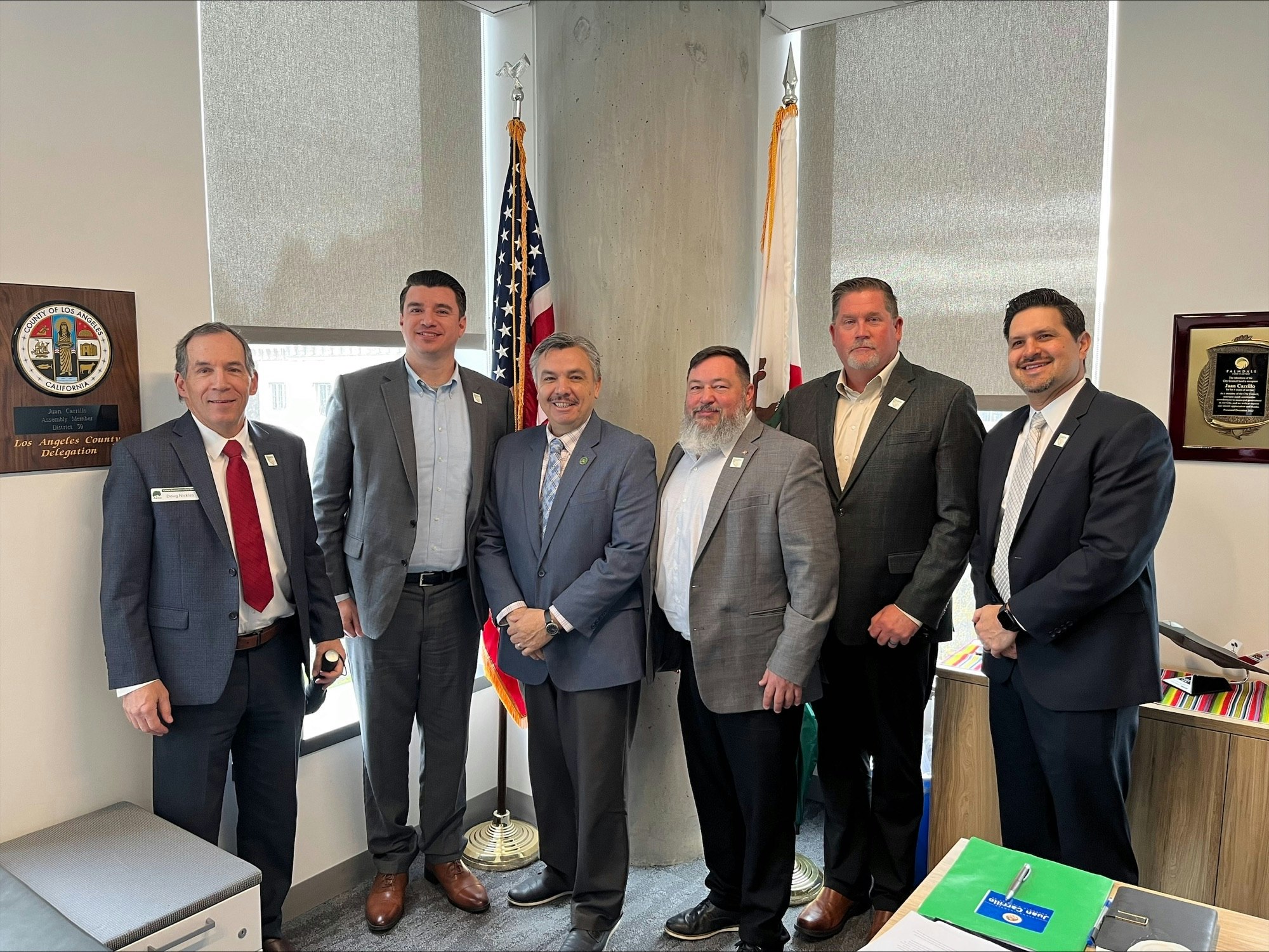 A group of six men in suits standing together in an office with U.S. flags and plaques on the walls.