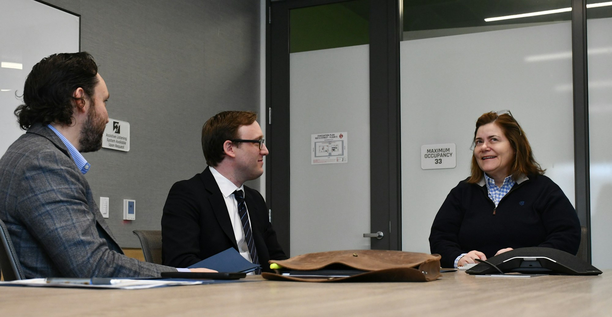 Three people in a meeting room engaged in conversation, with signs on the wall and items on the table.
