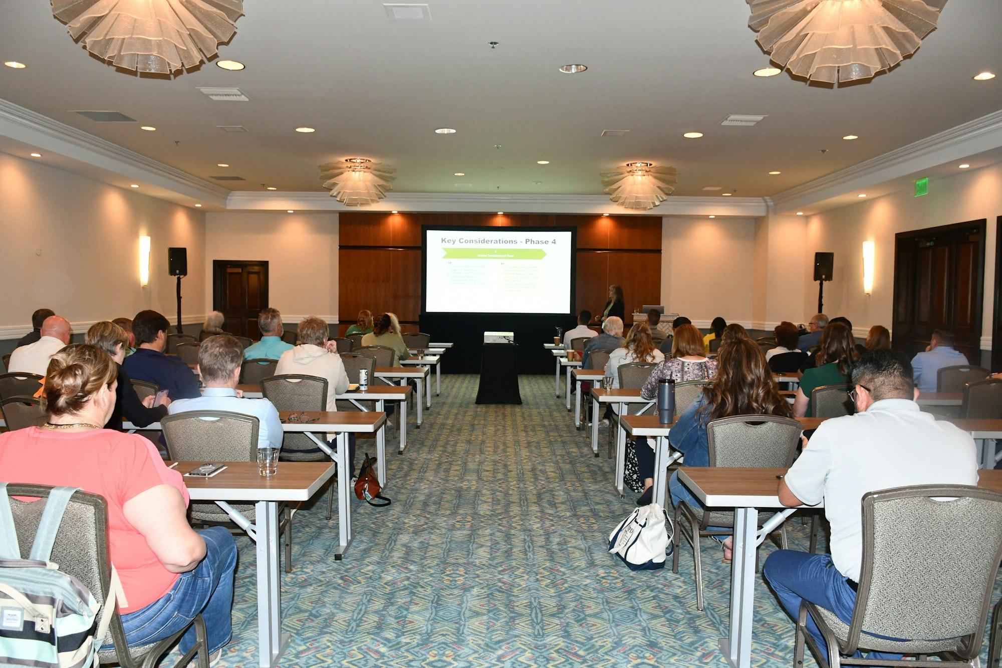 People attending a presentation in a conference room, seated at tables, facing a screen displaying "Key Considerations - Phase 4."