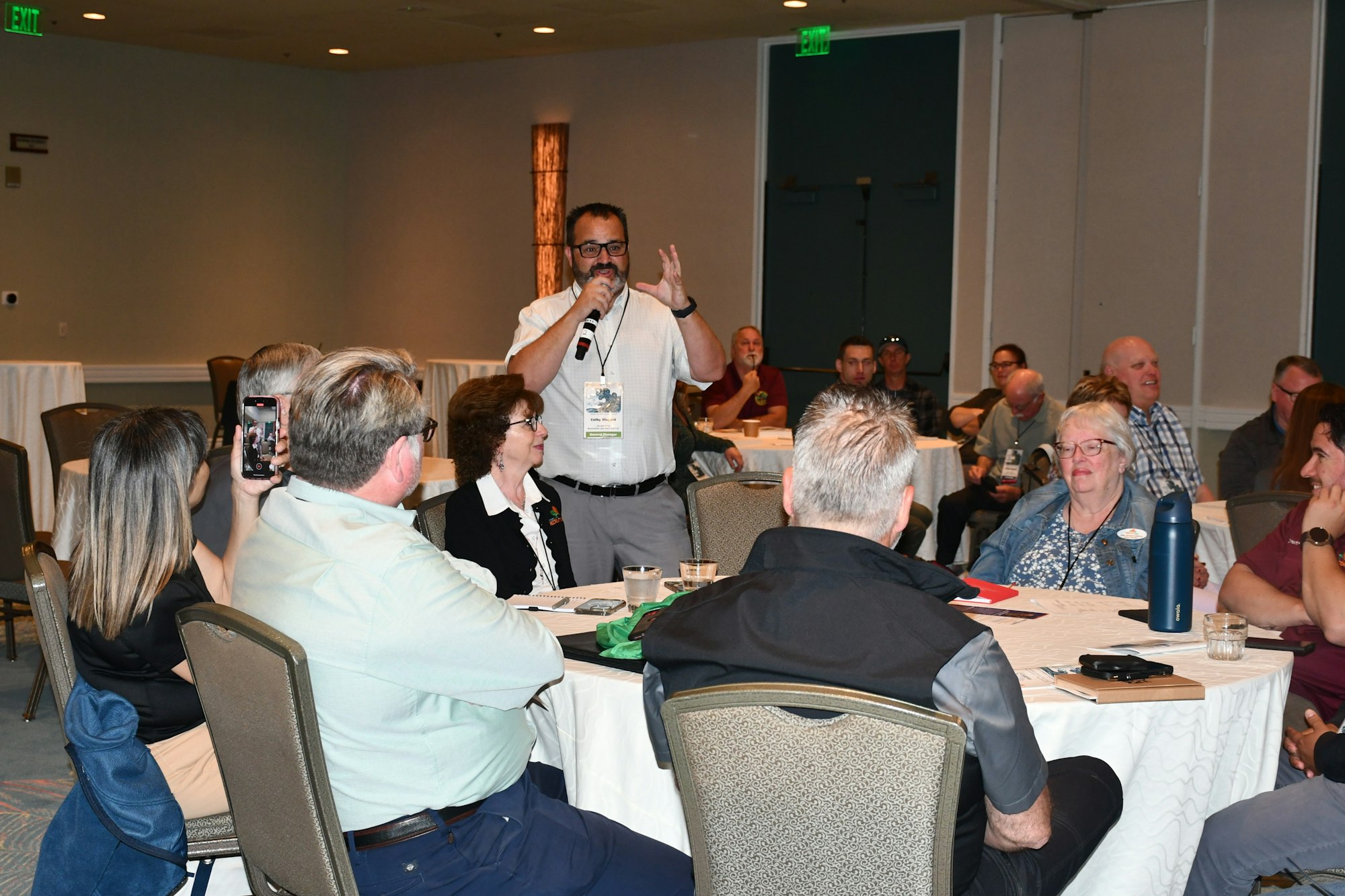 A man speaks with a microphone in a conference room, surrounded by seated attendees.