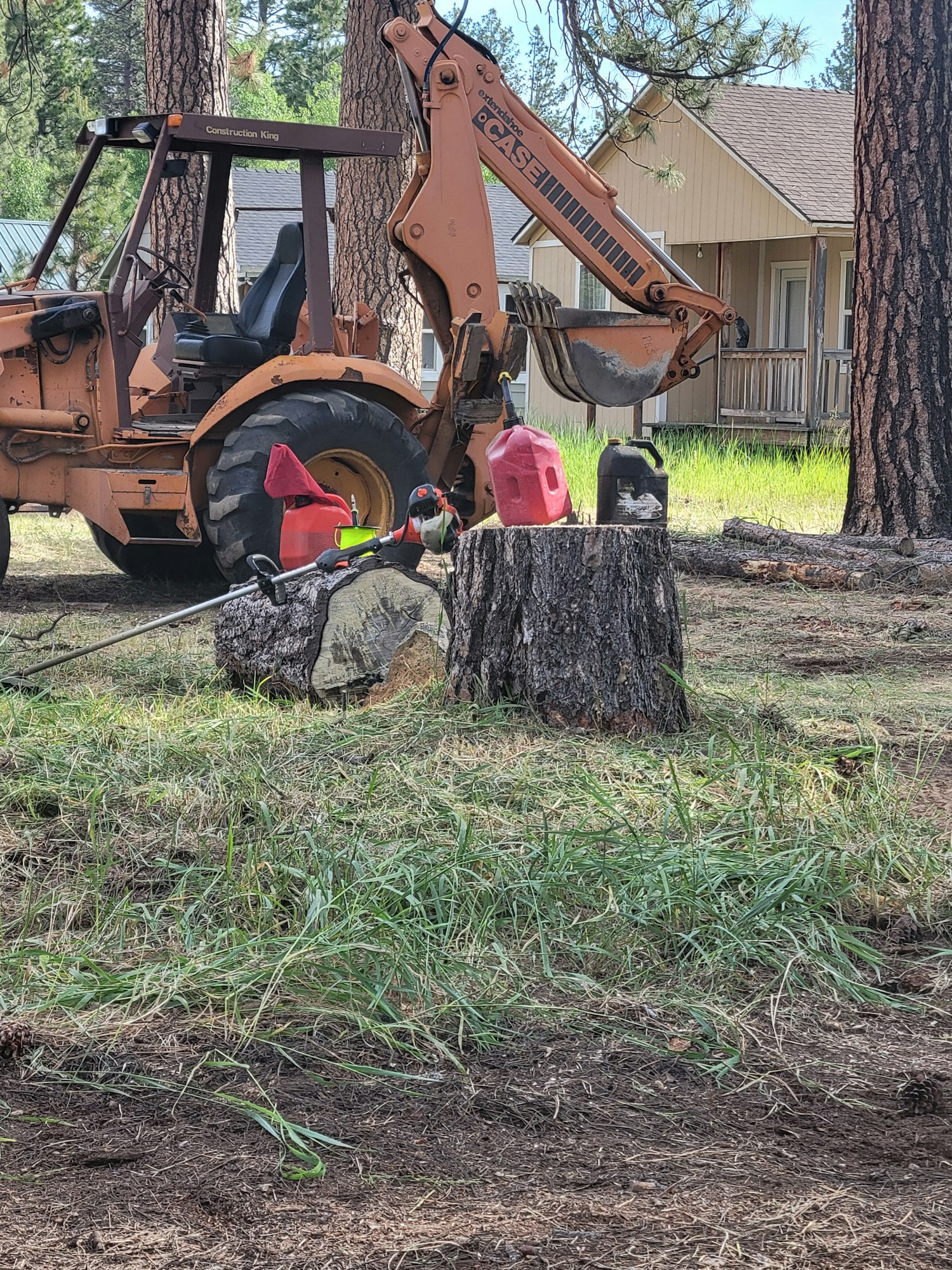 An excavator beside a tree stump, near a house, with safety cones and gas cans. Image is rotated 90 degrees.