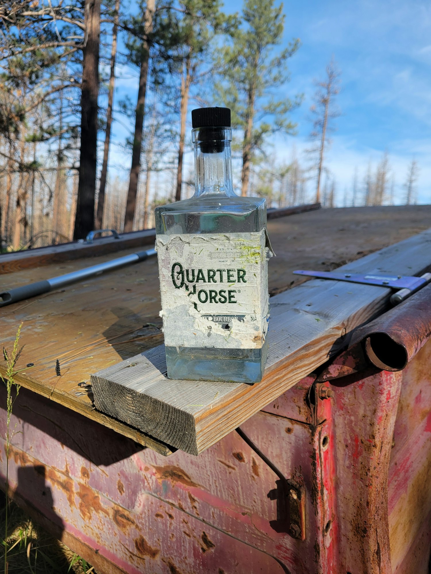 A glass bottle labeled "Quarter Horse" on a wooden surface with rusty red metal and trees in the background.