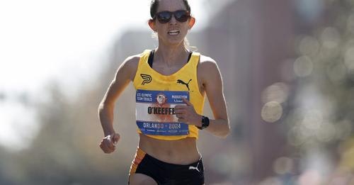 A female runner in a race, wearing a yellow top, black shorts, and sunglasses, with a race bib labeled "O'KEEFFE."