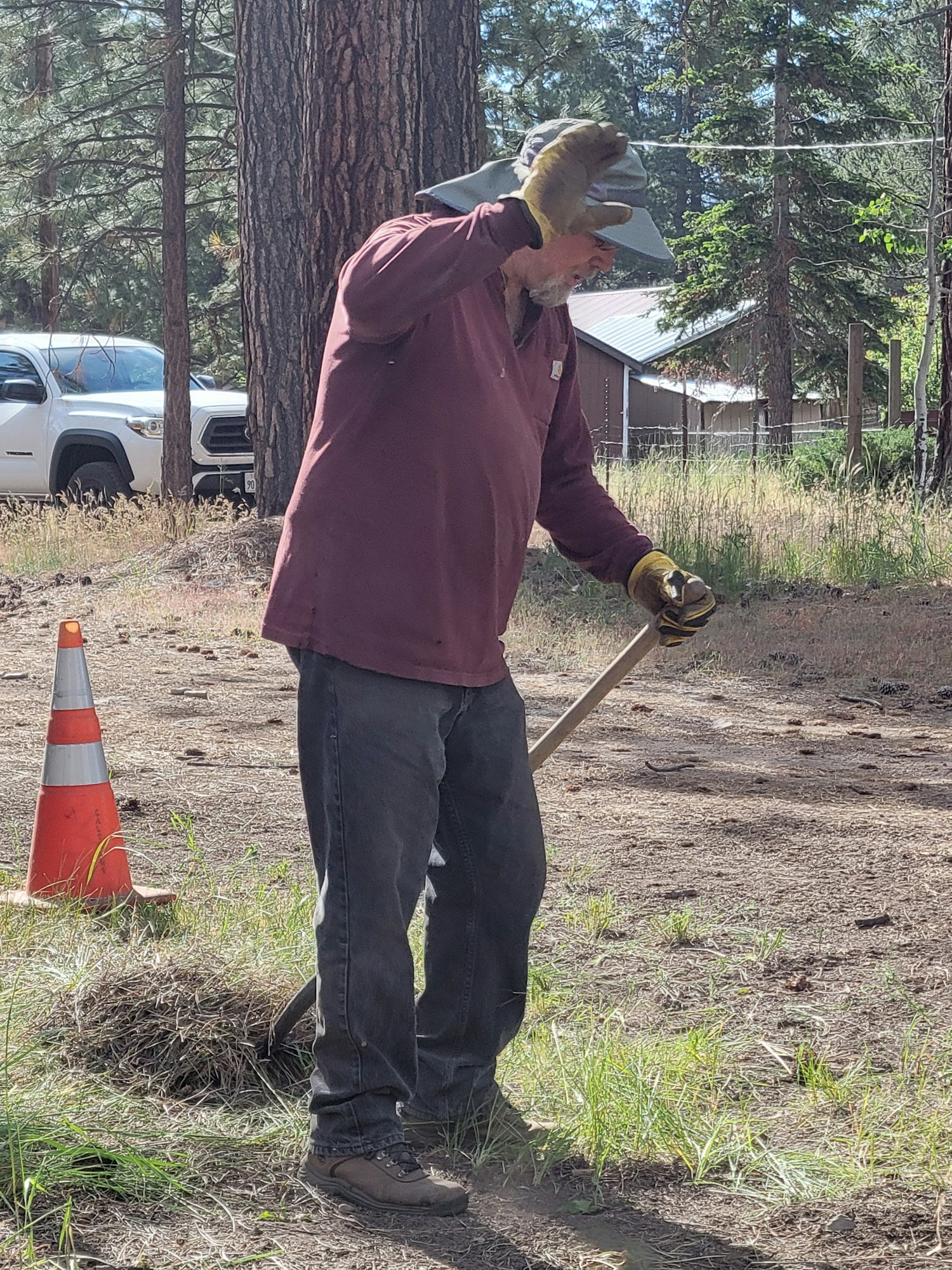 A man holding a shovel outdoors near trees and a traffic cone, with a white vehicle in the background. Image is rotated 90 degrees.