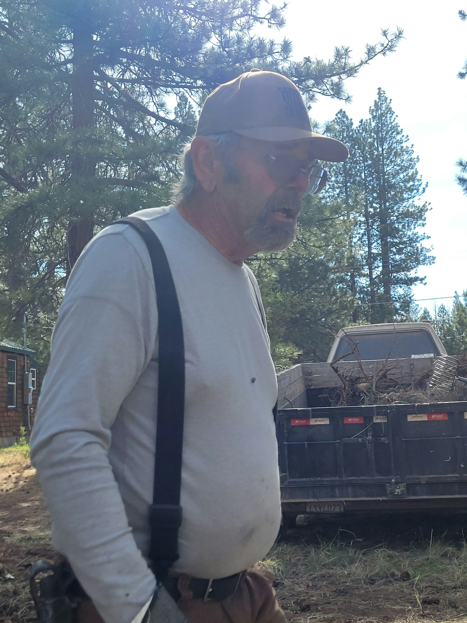 A man with a cap, suspenders, and a beard, standing outdoors next to a vehicle with trees in the background. Image is rotated.