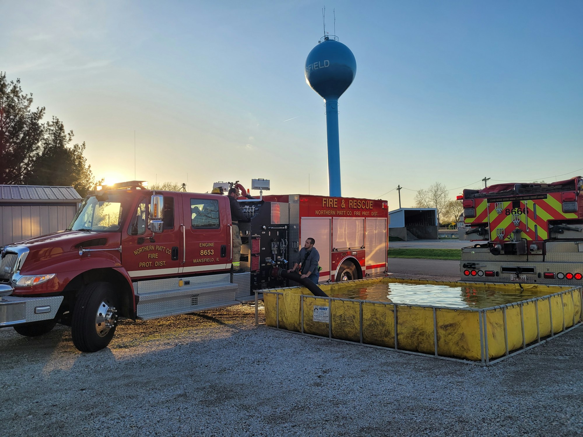 Two fire trucks near a water tank with a water tower in the background during sunset.